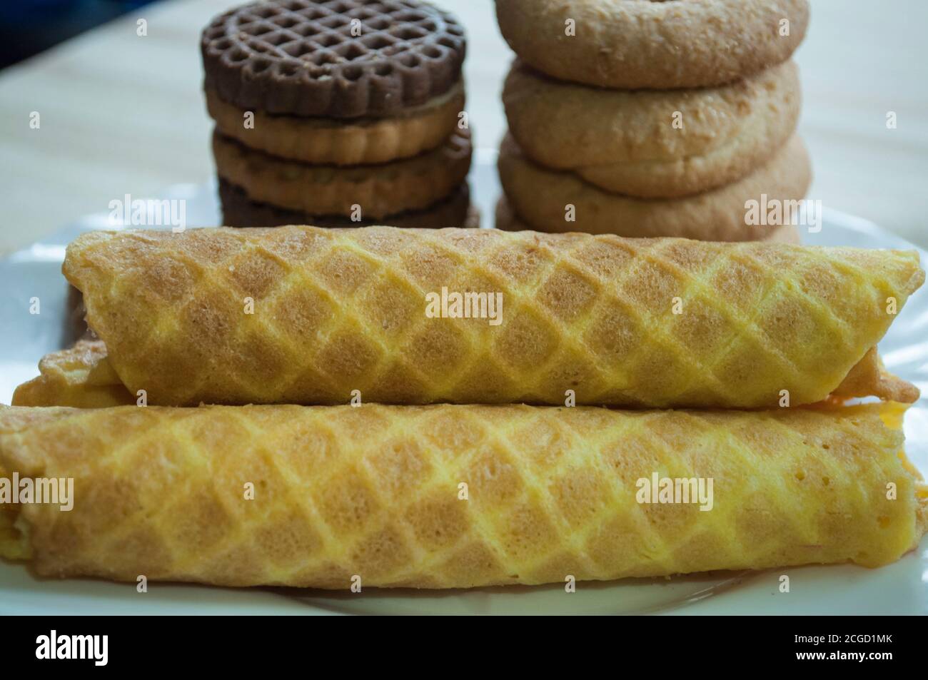 Waffle rolls and cookies on a white plate for tea, breakfast. Waffles in front. Composition on a wooden background. Visible waffle texture Stock Photo