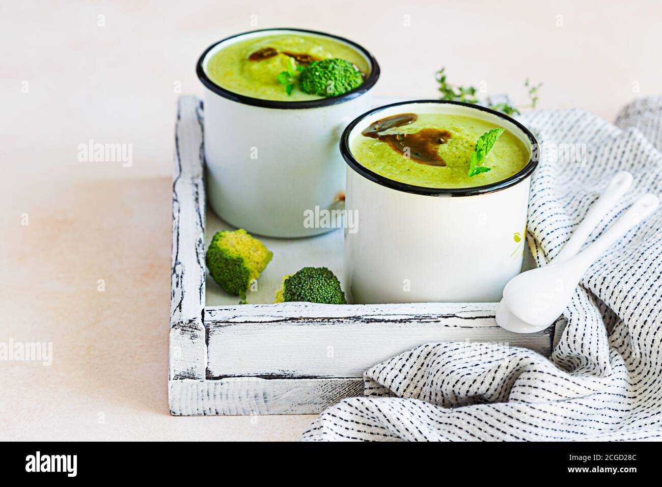 Two enamel mugs with healthy vegan broccoli soup with spicy oil and aromatic herbs over light stone background. Diet detox food concept. Stock Photo