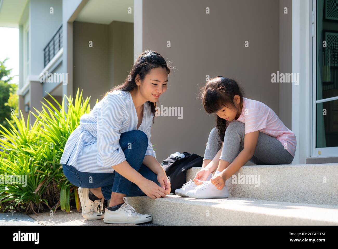 Asian mother help her daughter primary students in uniform to wearing their shoelace of shoes infront of home in the morning school routine for day in Stock Photo
