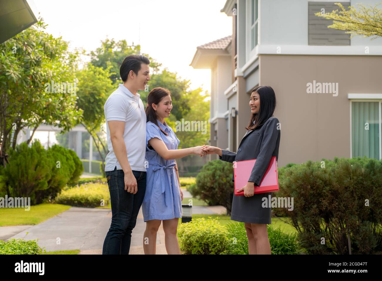 Happy Asian couple looking for their new house and shake hands with real estate broker after a deal. Young couple handshaking real estate agent after Stock Photo