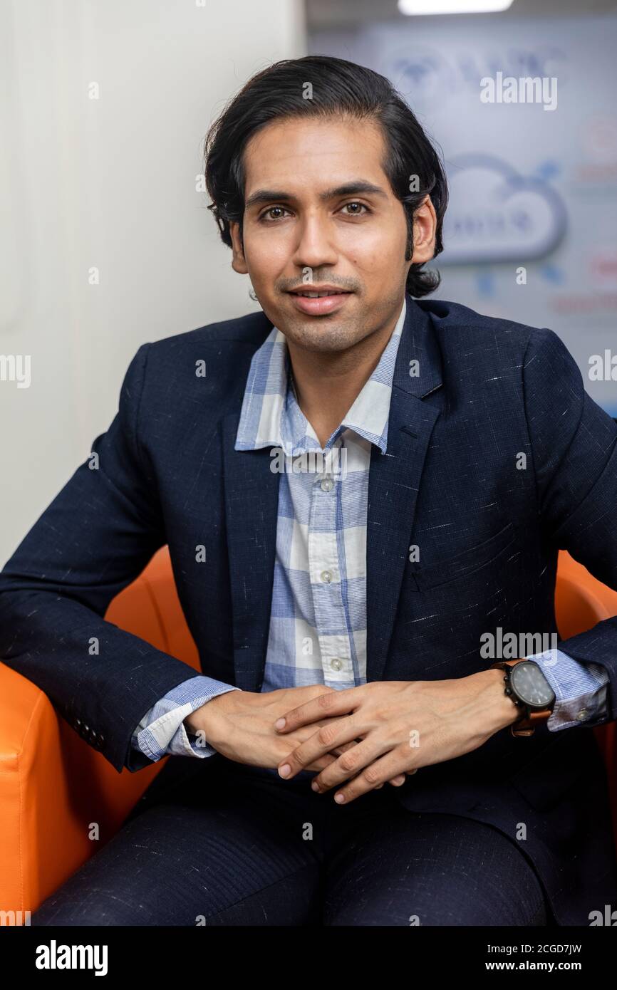 Portrait of young successful Indian business man in dark suit sitting on couch in a corporate environment, looking into camera. Stock Photo