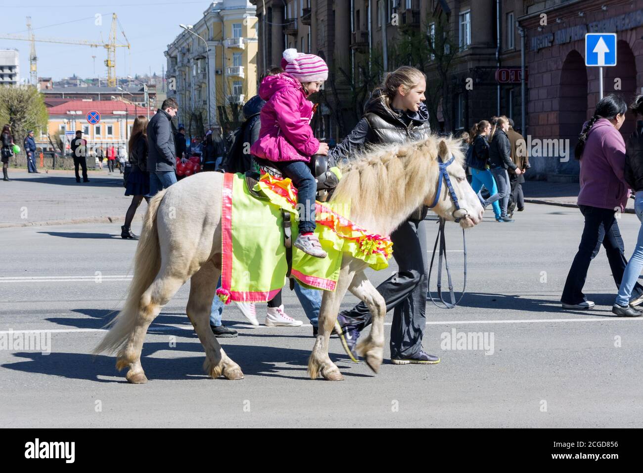 A little girl rides on a smart white pony driven by a girl along the avenue of Krasnoyarsk city during the celebration of Victory Day WWII. Stock Photo