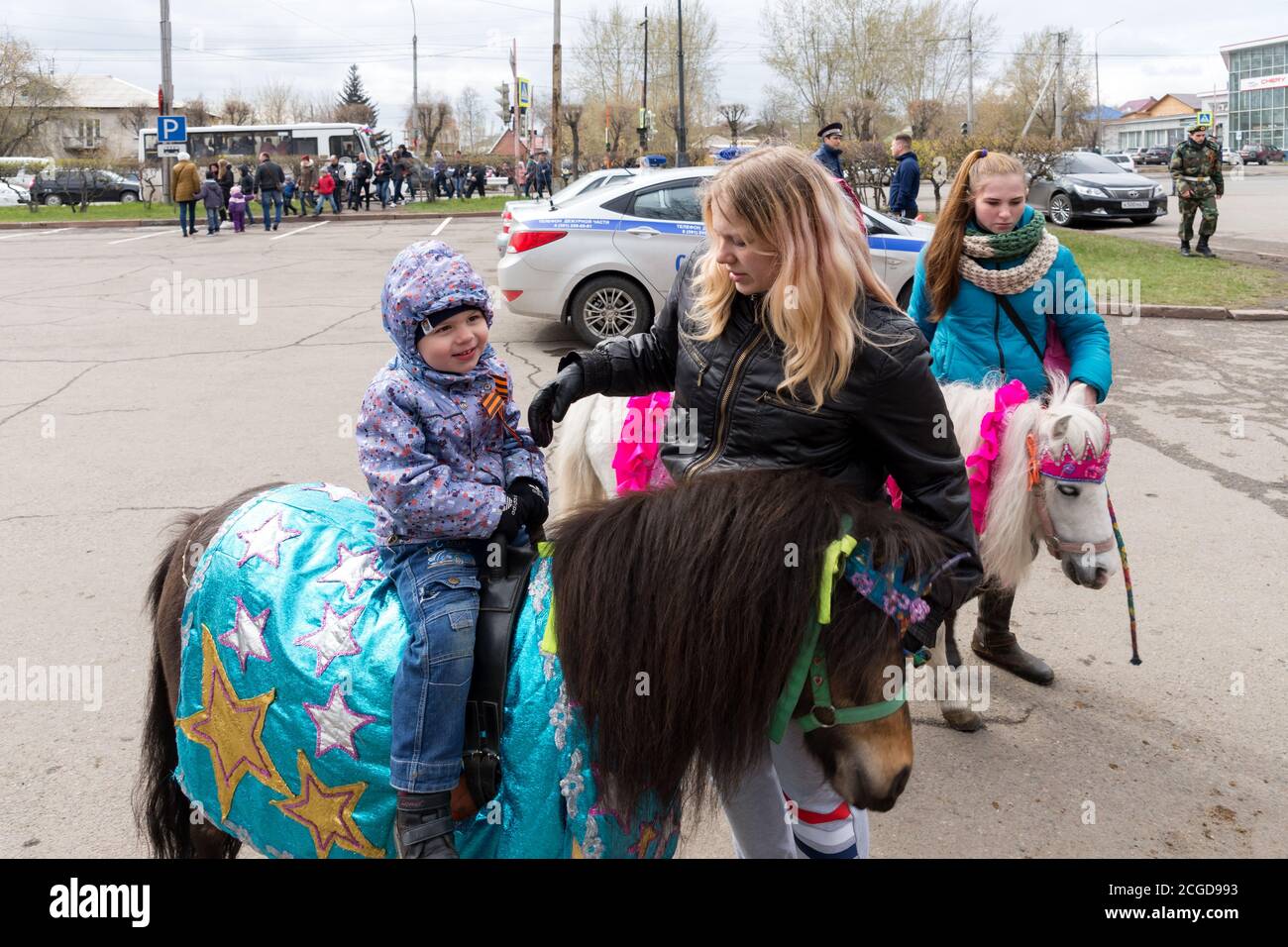 Little boy sits on a pony, a girl is standing next to her, against the background of a police car during the celebration of Victory Day WWII. Krasnoya Stock Photo