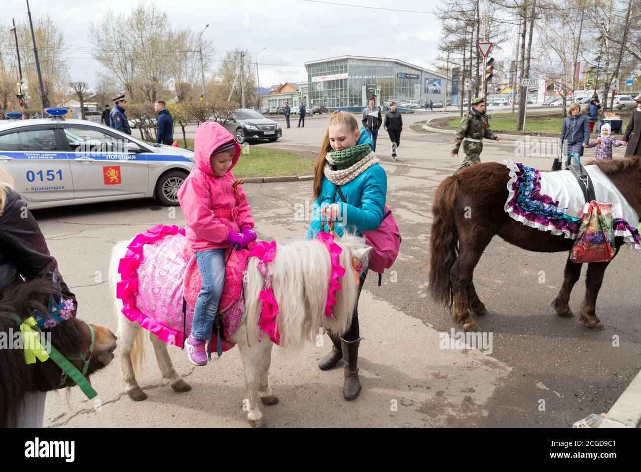 ittle girl sits on a pony, a girl is standing next to her, against the background of a police car during the celebration of Victory Day WWII Stock Photo