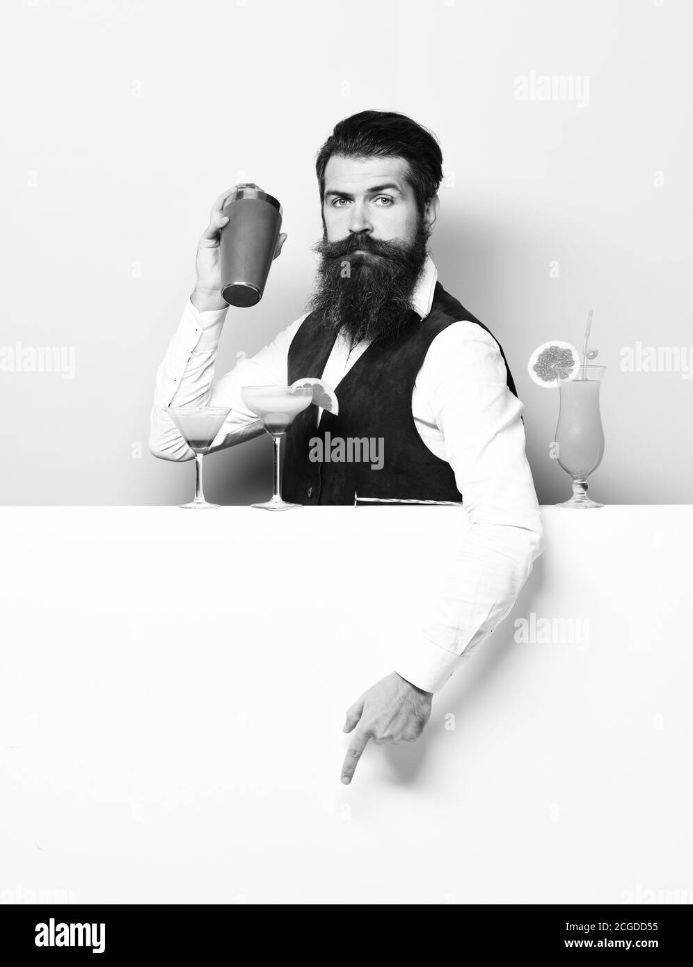handsome bearded barman with long beard and mustache has stylish hair on serious face holding shaker and made alcoholic cocktail in vintage suede leather waistcoat on purple green studio background Stock Photo