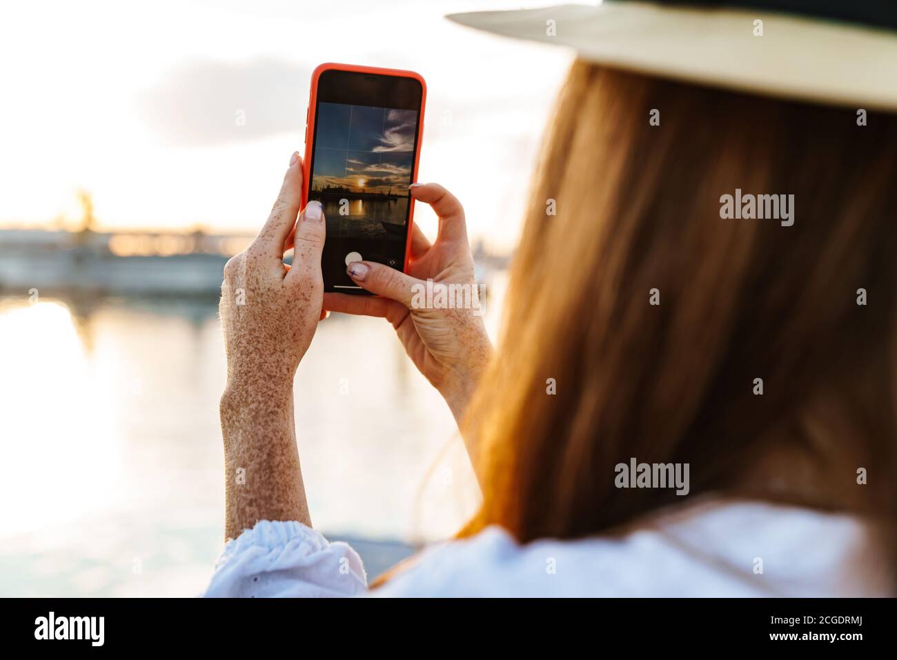 Image of young ginger woman taking photo of sunset on mobile phone at promenade Stock Photo