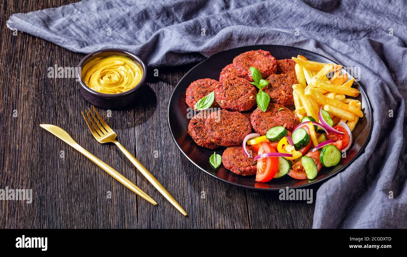 Vegan beetroot, mushrooms, black bean patties served with french fries and tomato, cucumber red onion salad on a black plate with nutritional yeast sa Stock Photo