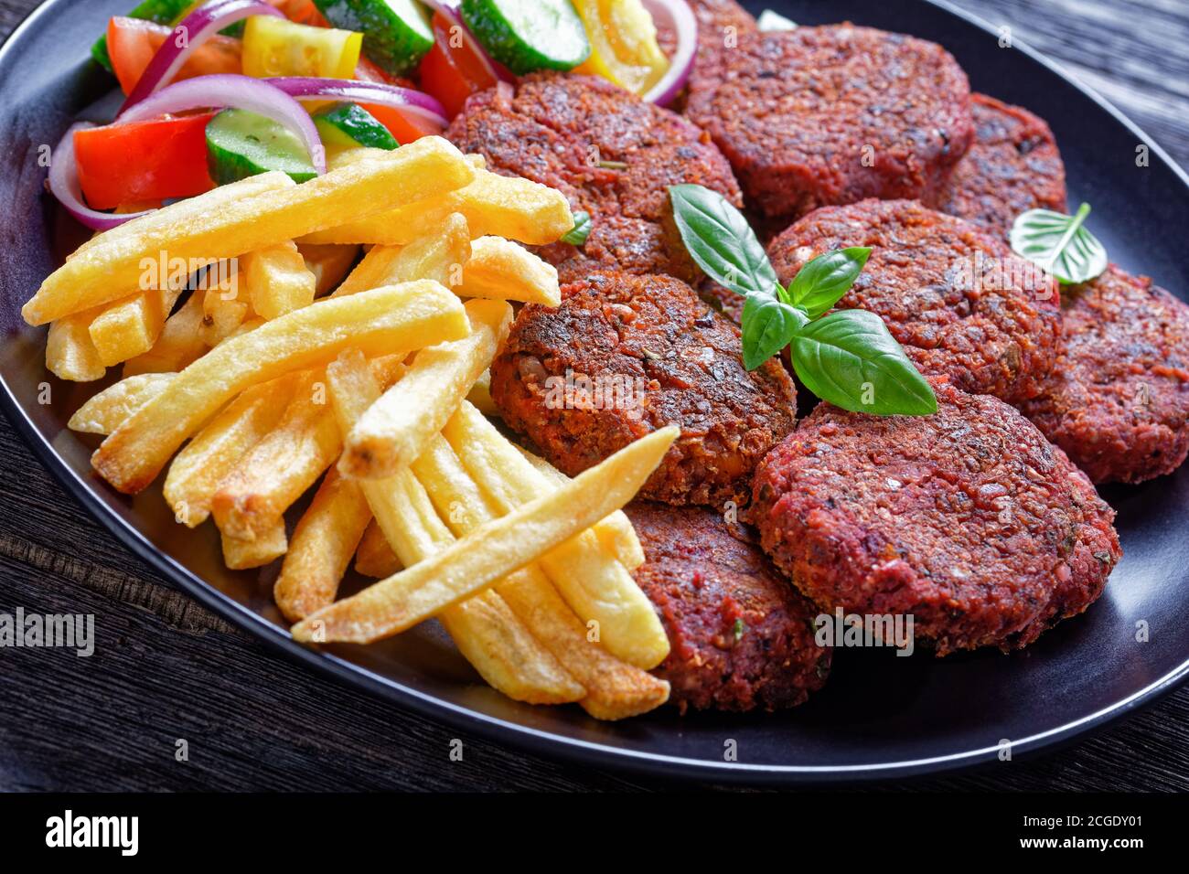French fry, fresh salad of tomato cucumber and red onion with fresh basil served with beetroot patties of mushrooms and black beans on a black plate o Stock Photo