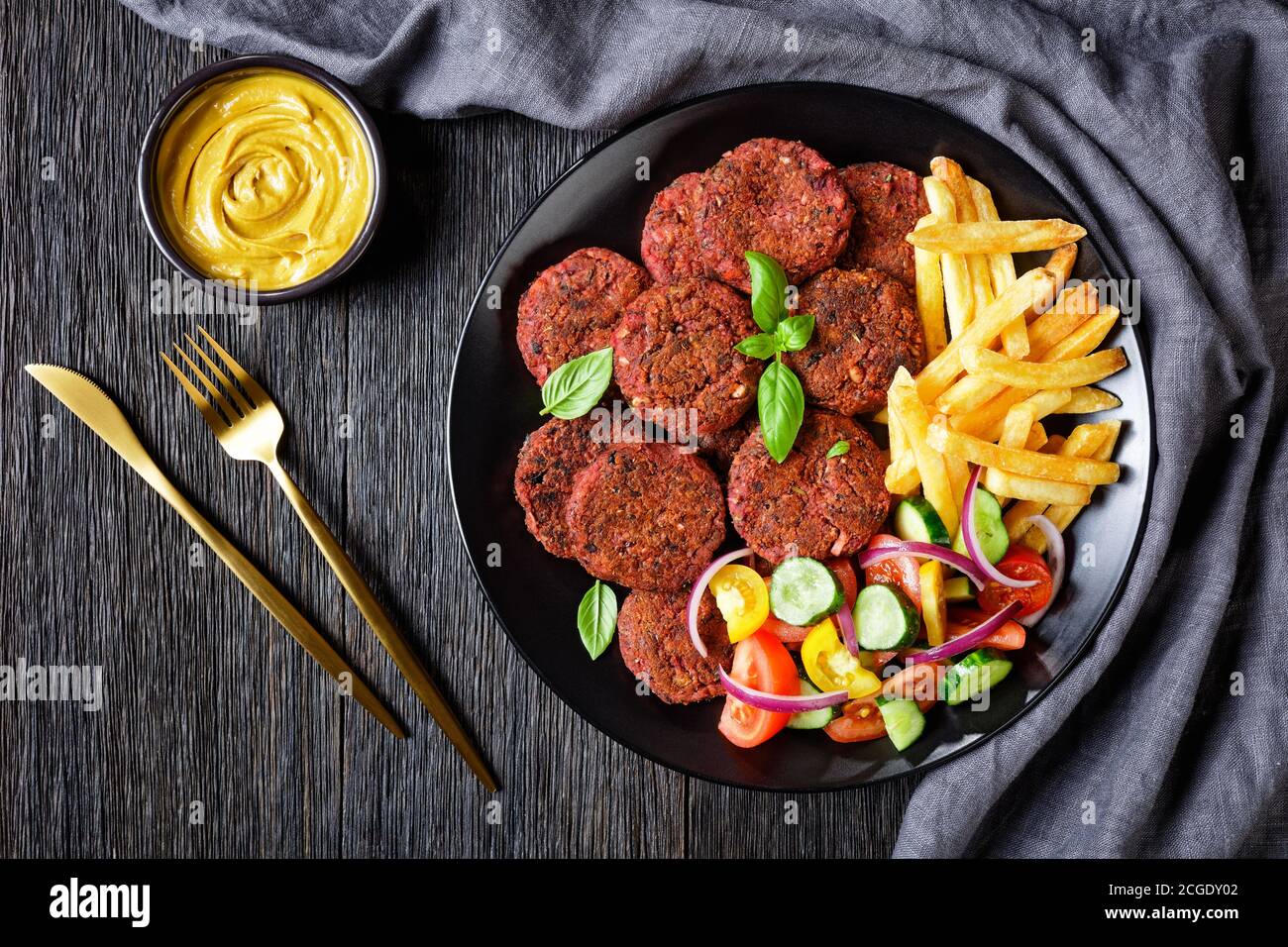 Vegan beetroot cakes of mushrooms, black bean served with french fries and fresh vegetable salad on a black plate with nutritional yeast sauce on a da Stock Photo