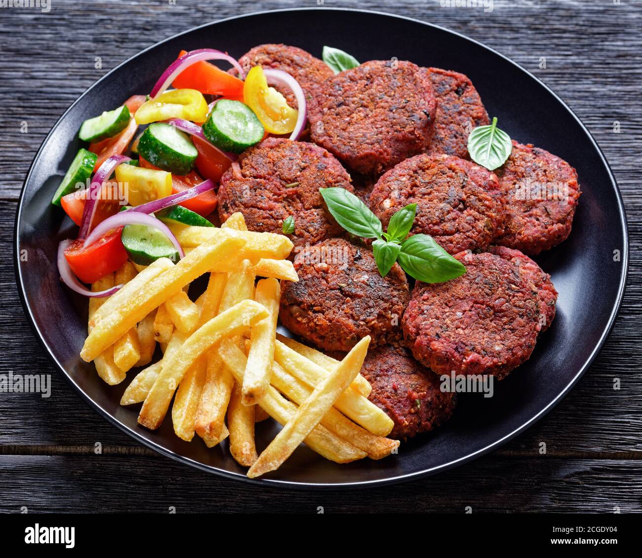 Roasted beet burger patties with french fry, fresh salad of tomato cucumber and red onion with fresh basil served on a black plate on a dark wooden ta Stock Photo