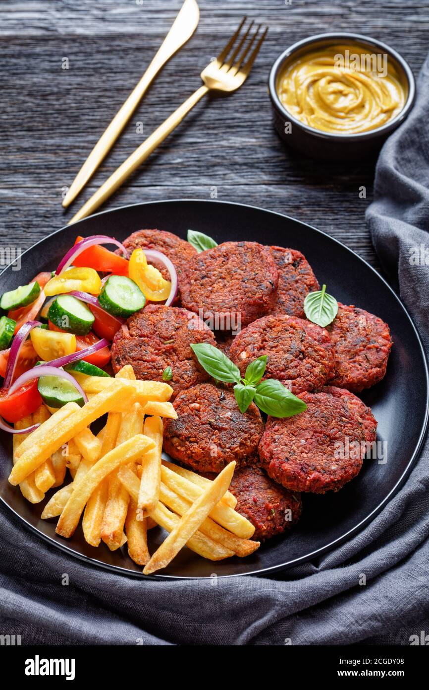 Vegan beetroot patties of mushrooms, black bean served with french fries and fresh vegetable salad on a black plate with vegan cheese sauce on a dark Stock Photo