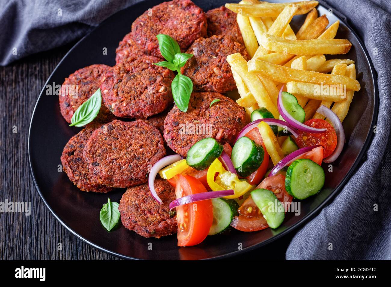 Vegan meal: beetroot cakes of mushrooms and black bean served with french fries and vegetable salad of tomato, cucumber and red onion on a black plate Stock Photo