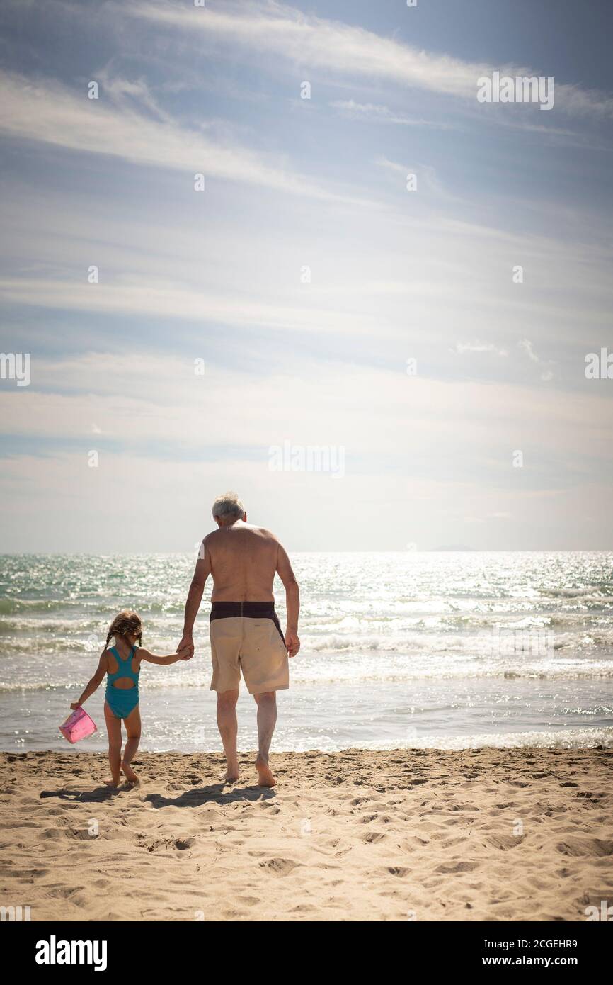 Caucasian grandfather and granddaughter walking hand in hand on the beach. Back view, vertical shot. Stock Photo