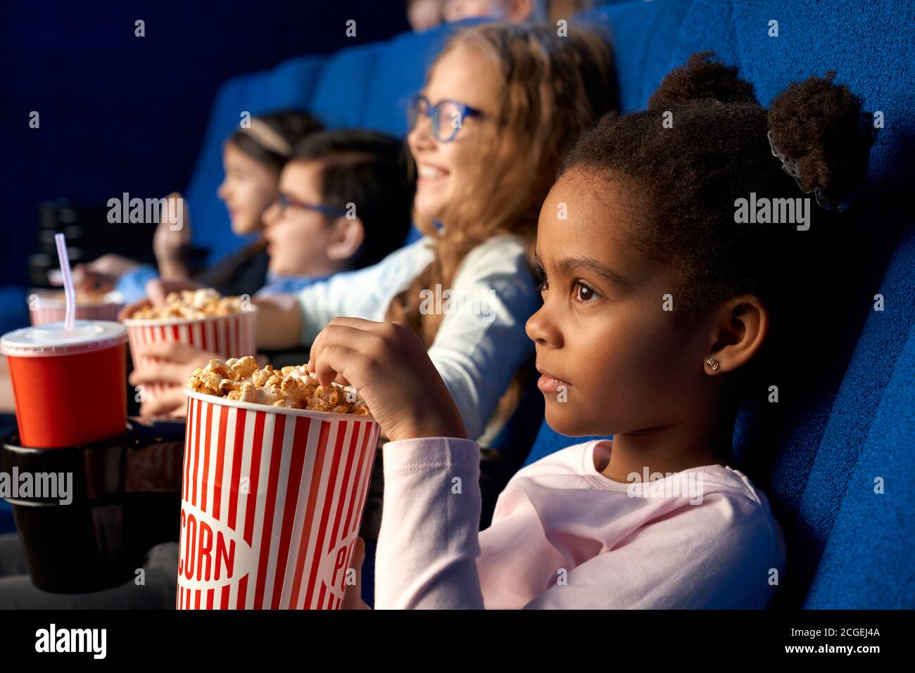Adorable little female kid sitting with friends, eating popcorn and enjoying time. Beautiful concentrated african girl with funny hairstyle watching movie in cinema. Concept of entertainment, leisure. Stock Photo