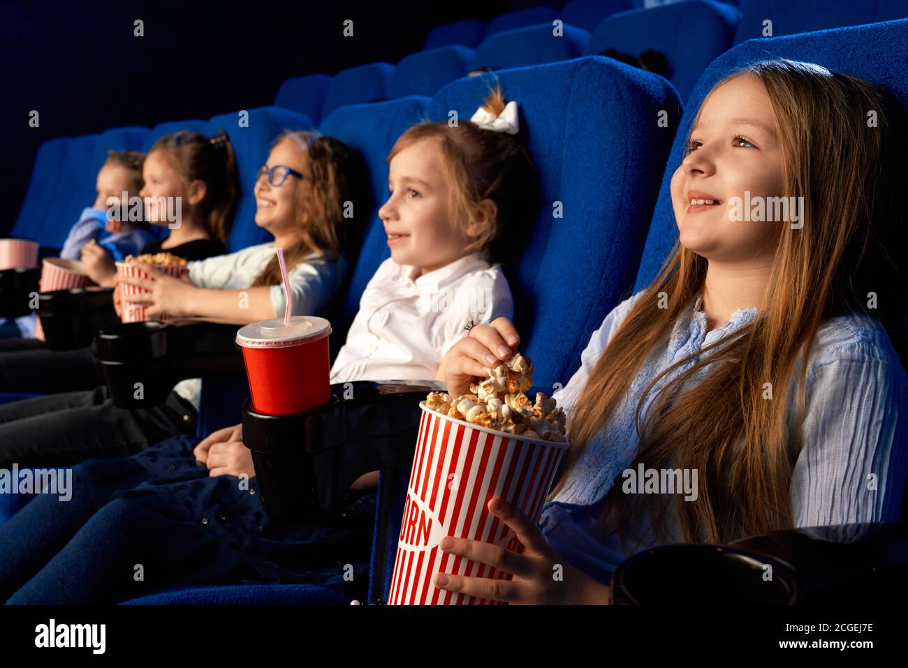 Selective focus of smiling little girl holding popcorn bucket, sitting with laughing friends in comfortable chairs in cinema. Children watching cartoon or movie, enjoying time. Entertainment concept. Stock Photo