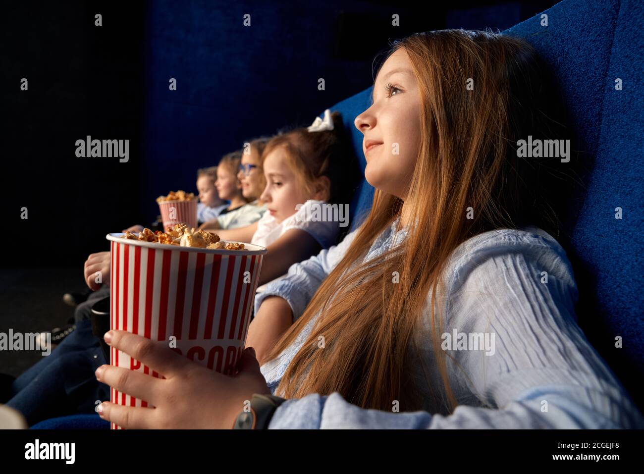Selective focus of pretty little girl holding popcorn bucket, sitting with friends in comfortable chairs in cinema. Children watching cartoon or movie, having fun. Leisure, entertainment concept. Stock Photo