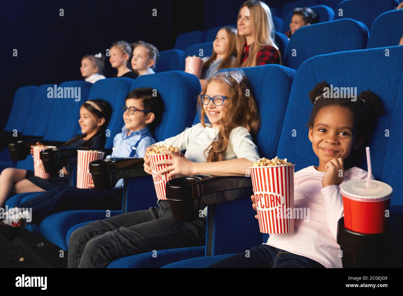 Lovely girl sitting in cinema with friends, looking at camera and smiling while watching movie. Little adorable african female kid eating popcorn and drinking sweet water. Cocept of entertainment. Stock Photo