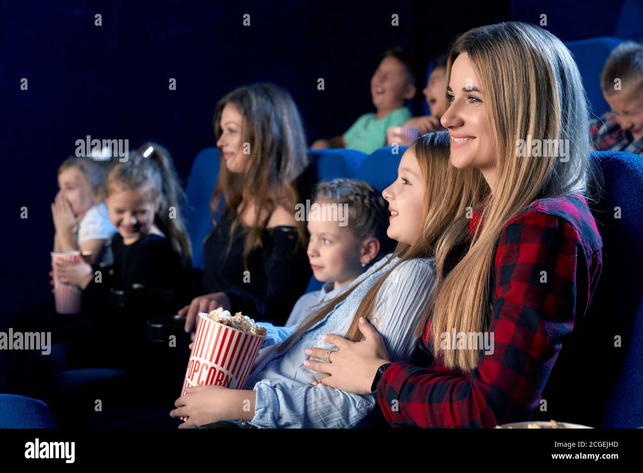 Side view of family spending time together in cinema. Attractive young mother holding little daughter on knees and smiling while watching film and eating popcorn. Concept of leisure, entertainment. Stock Photo