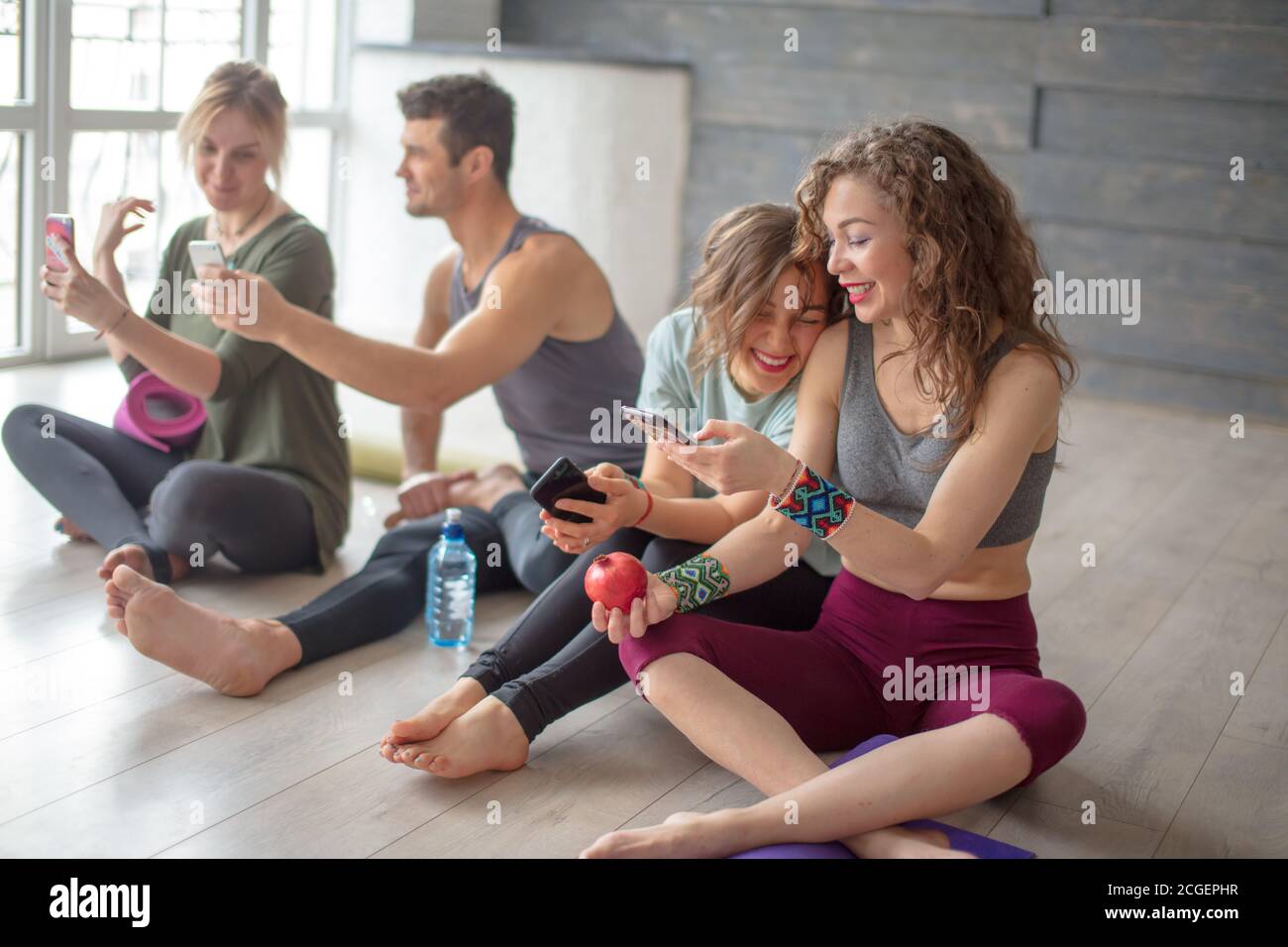 Group of female and male yoga pupils relaxing with fresh fruit and water after yoga training at health club. Friends checking messages and socialmedia Stock Photo