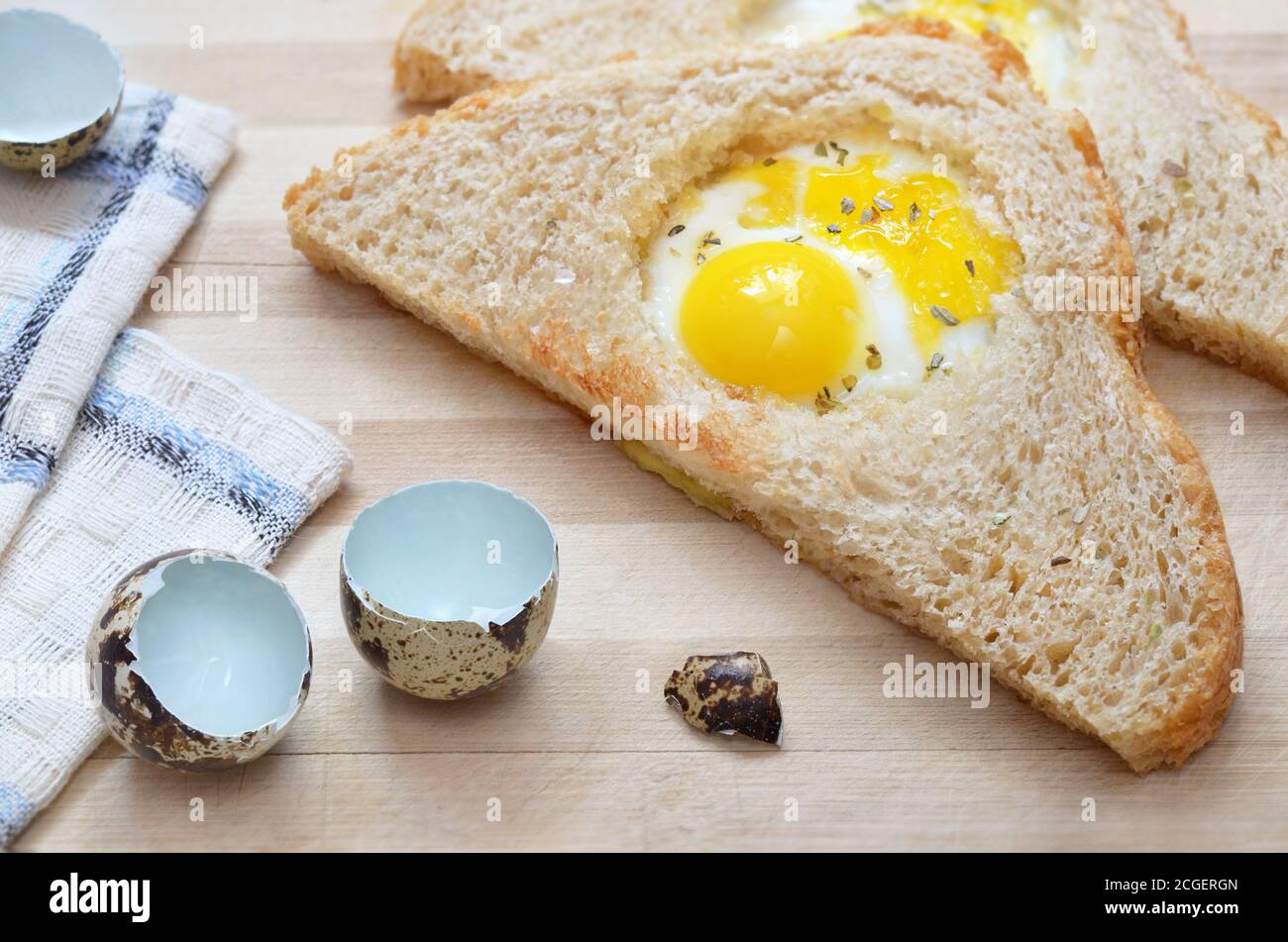 Fried slice of bread with quail egg on a wooden cutting board with a kitchen towel and eggshells, selective focus. Healthy eating concept. Stock Photo