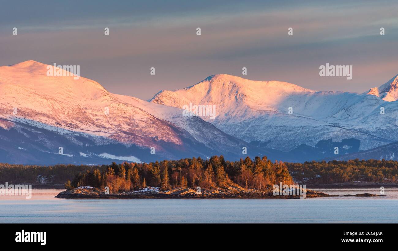 Panoramic vista of the coast of Norway with snow covered mountains and small island with trees in beautiful soft evening light Stock Photo