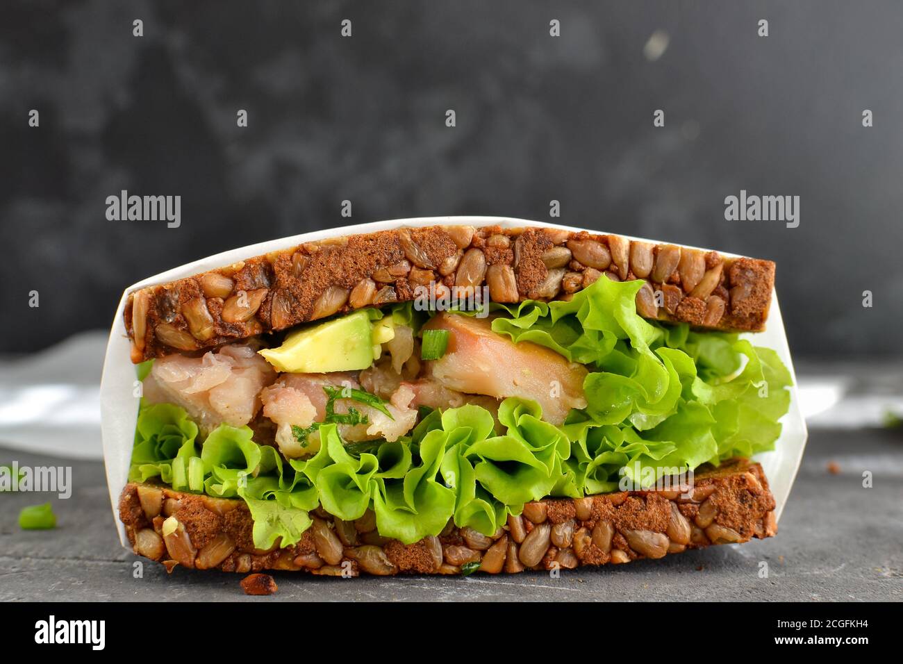 Dark bread sandwich with seeds. Sandwich with lettuce, avocado and fish. Healthy lunch or brunch. Dark background. Close-up. Stock Photo