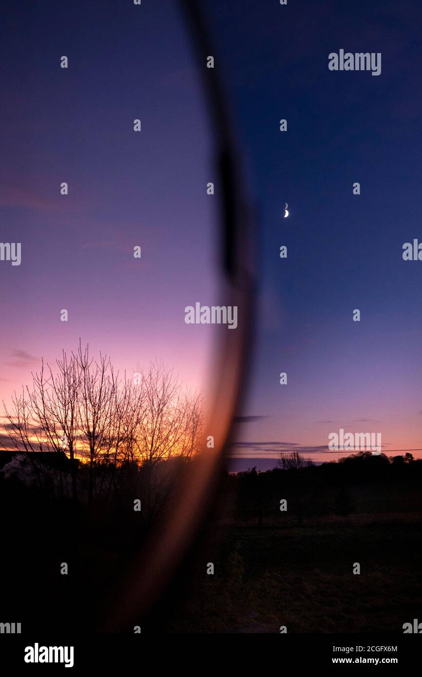 Looking out of a window at the deeply coloured sky at sunset reflected in a mirror with a bright moon in the distance. Stock Photo