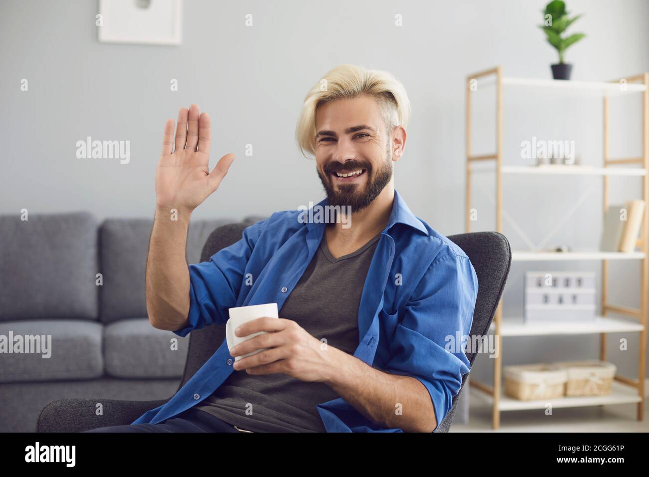 Smiling man sitting at home and greeting somebody with hand online looking at web camera Stock Photo
