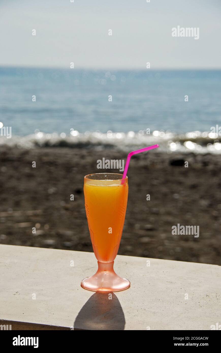 Glass of orange juice with straw with the beach and sea to the rear, Lagos, Malaga Province, Andalusia, Spain, Western Europe. Stock Photo