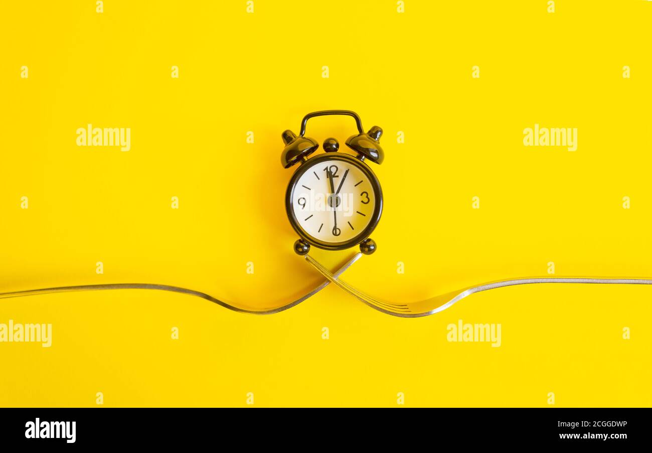 Black clock on a yellow background. Alarm clock on two forks, trend concept time . Stock Photo