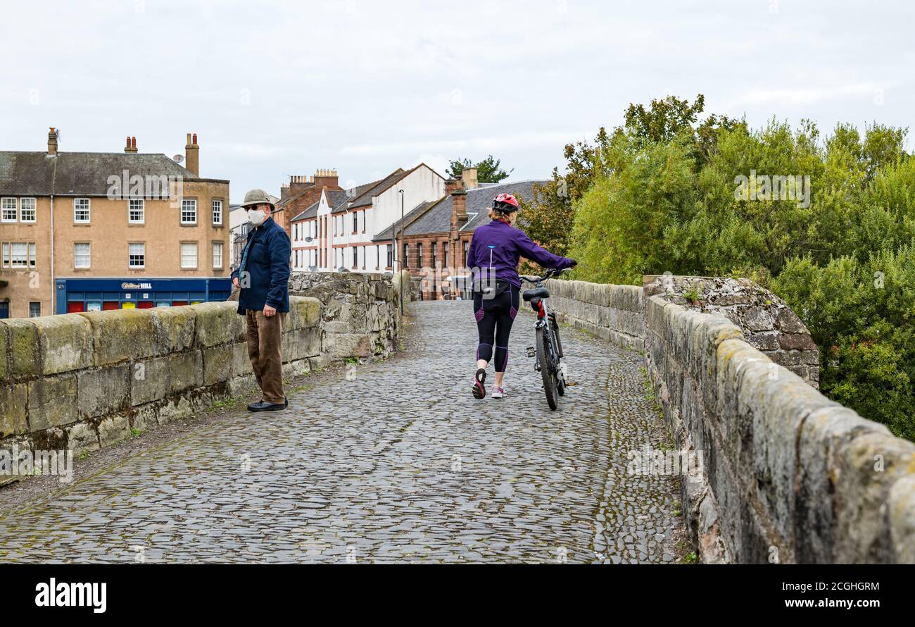 Older man wearing face mask and woman pushing bike social distancing on Roman bridge, Musselburgh, East Lothian, Scotland, UK Stock Photo