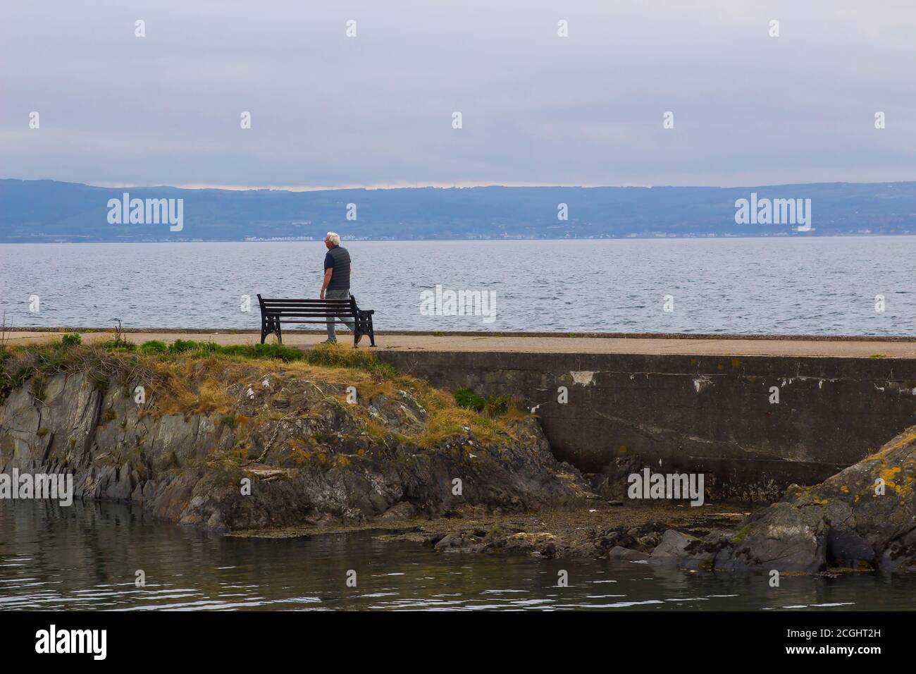 17 June 2020 A lone elderly man walks along the concrete wall that is part of the ancient Long Hole Harbour in Bangor County Down Northern Ireland on Stock Photo