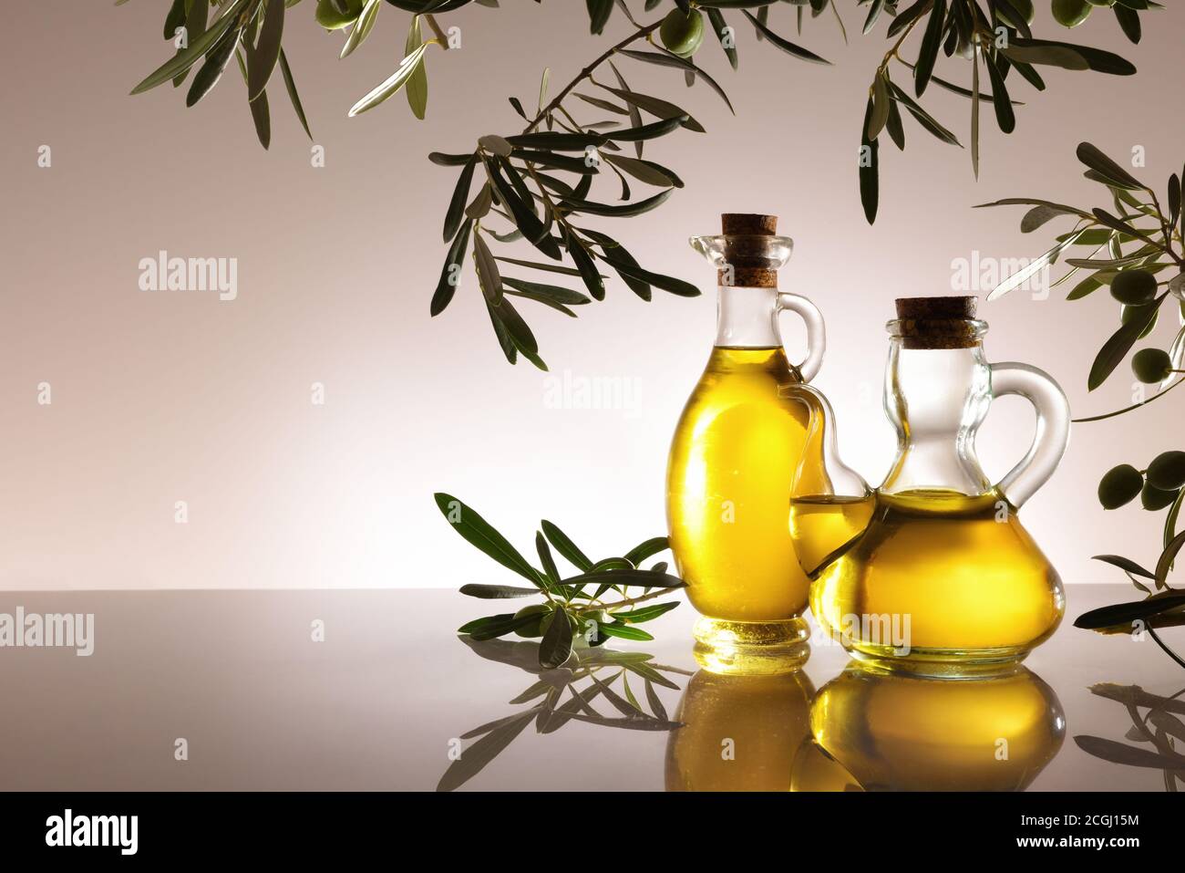 Glass containers full with olive oil backlit on glass table and gray gradient isolated background with olive branches. Front view. Stock Photo
