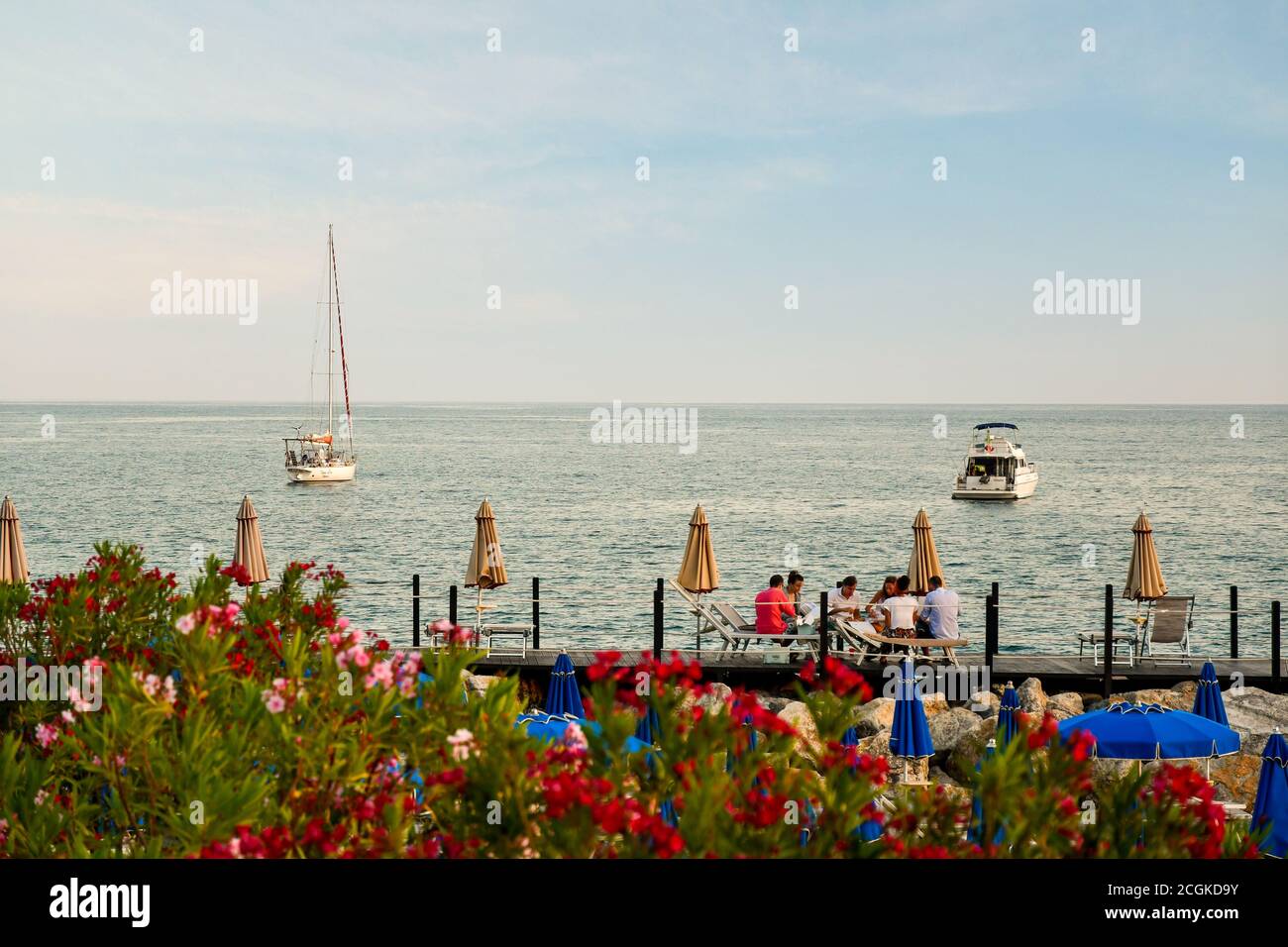 Group of young people having dinner sitting on deck chairs on a wooden pier on the sea shore with flowering oleander trees, Lerici, La Spezia, Italy Stock Photo