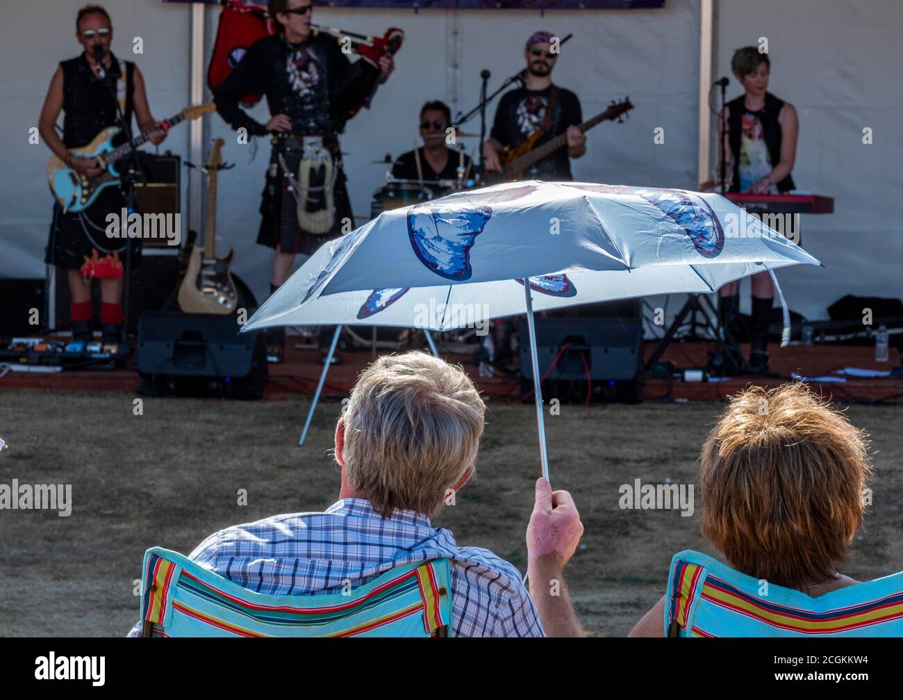 a middle aged couple under a parasol shading from the summer sun watching a band or group performing live on stage at a festival. Stock Photo