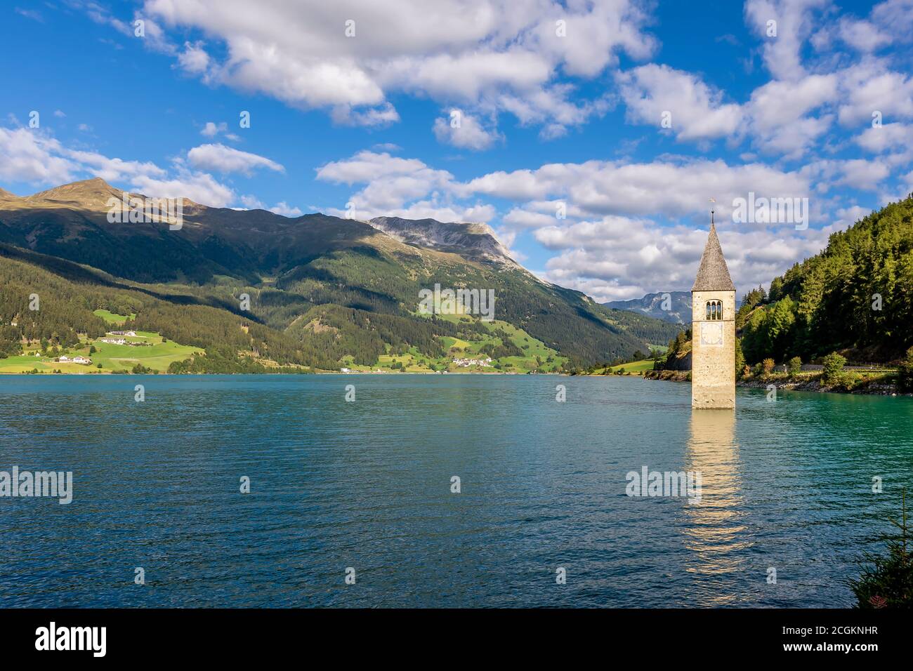 The ancient submerged bell tower of Lake Resia in Val Venosta, South Tyrol, Italy, emerges from the water against a beautiful blue sky with white clou Stock Photo