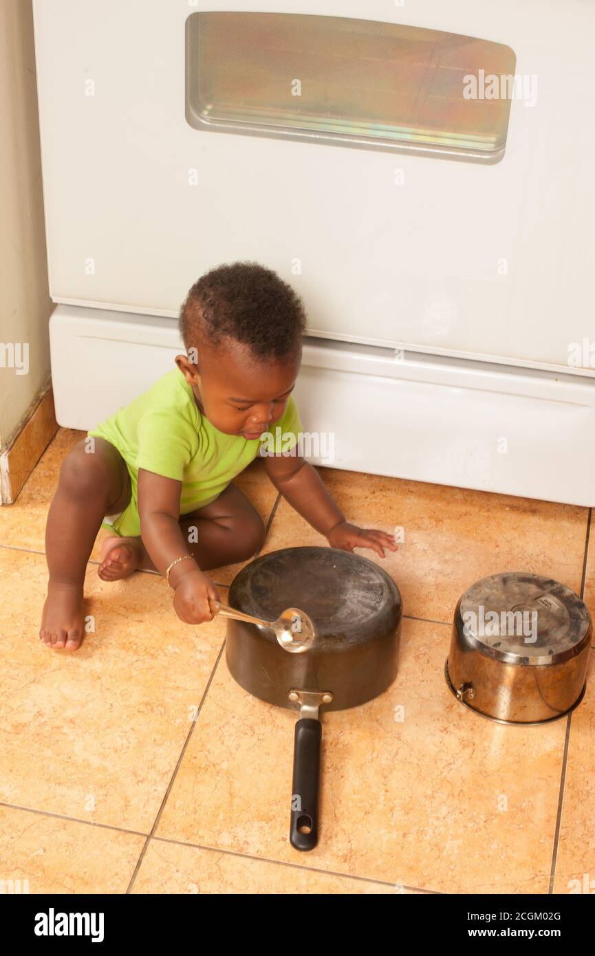 12 month old baby boy exploring sound hitting pot with metal spoon on floor in kitchen African American Stock Photo