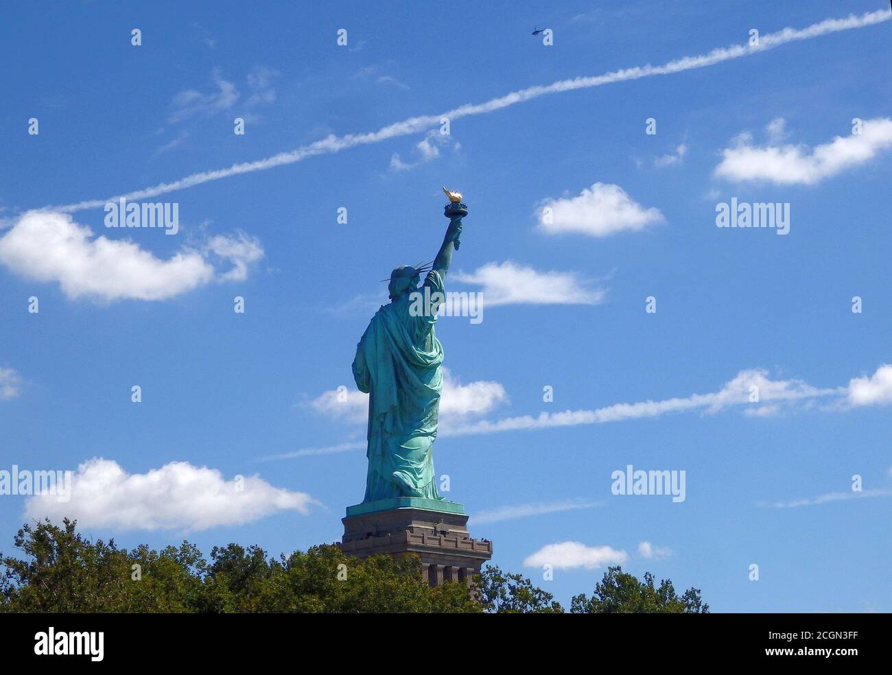 The back of the Statue of Liberty National Monument with a contrail in the sky and a helicopter passing over, New York City, United States Stock Photo