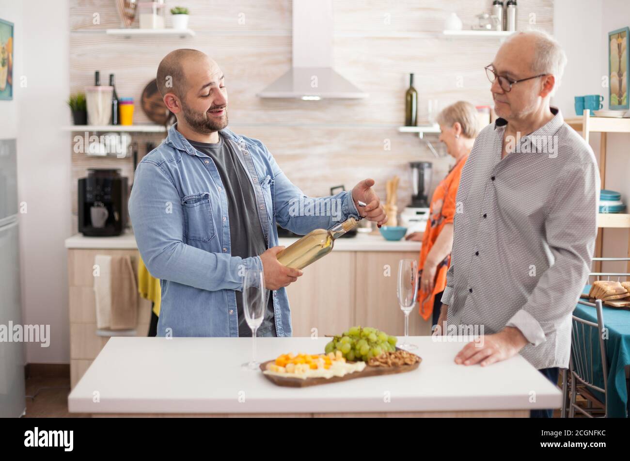 Son holding wine bottle in kitchen while having a conversation with his senior old. Wfe and mother preparing delicous lunch for family meeting. Stock Photo