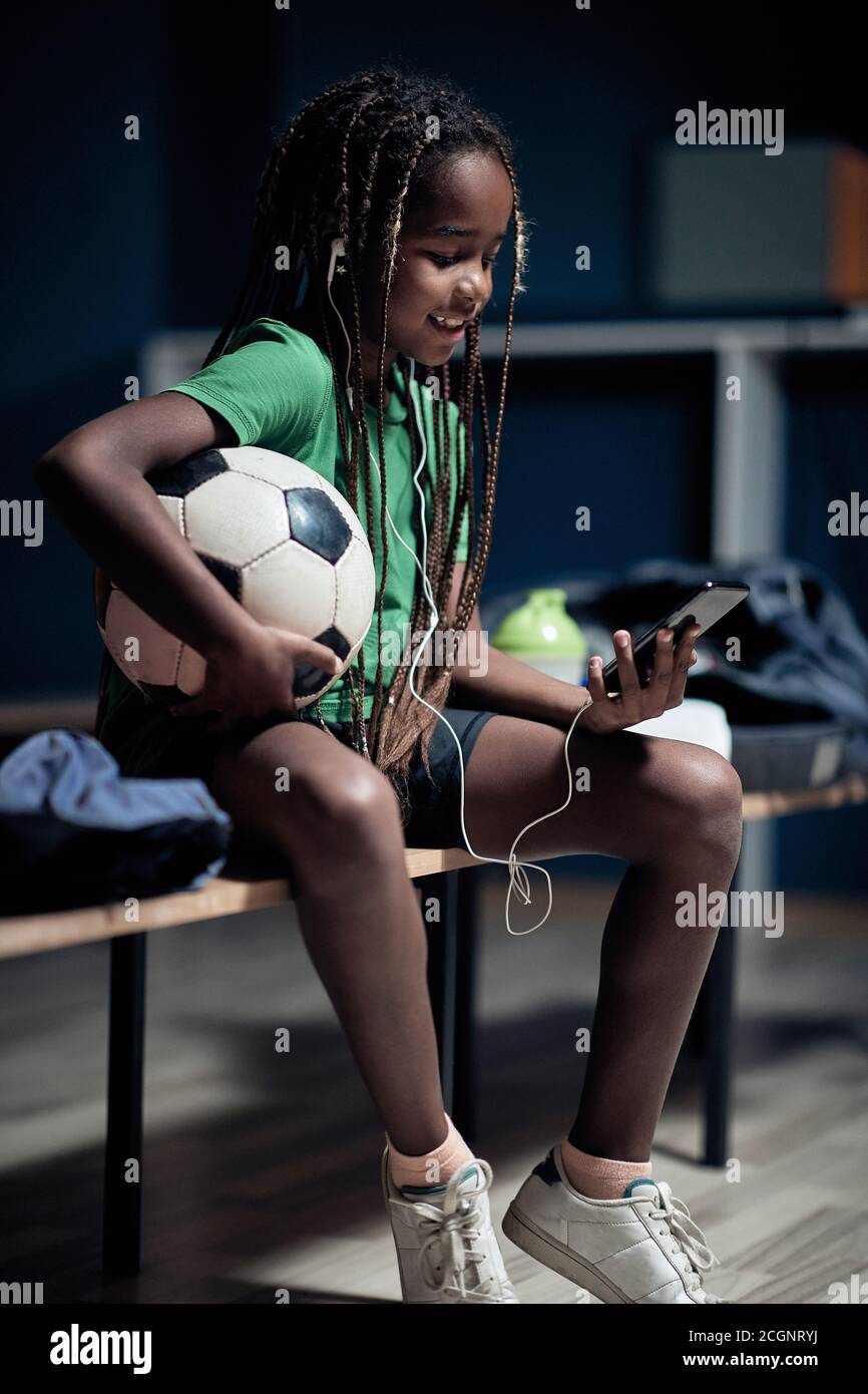 A little soccer player enjoying content on cell phone waiting for a training  in a locker room Stock Photo