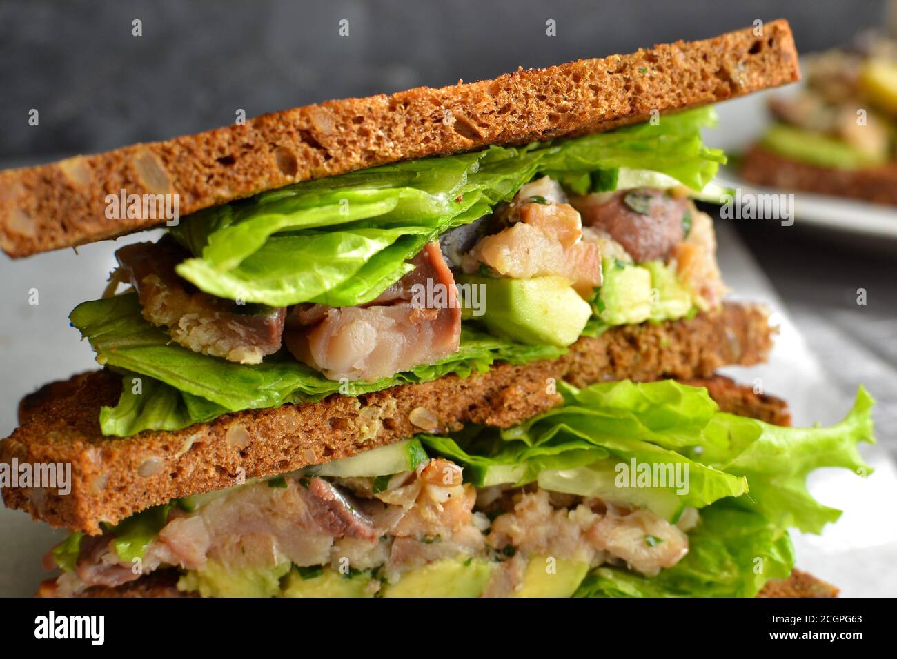 Dark bread sandwich with seeds. Sandwich with lettuce, avocado and fish. Healthy lunch or brunch. Dark background. Close-up. Stock Photo