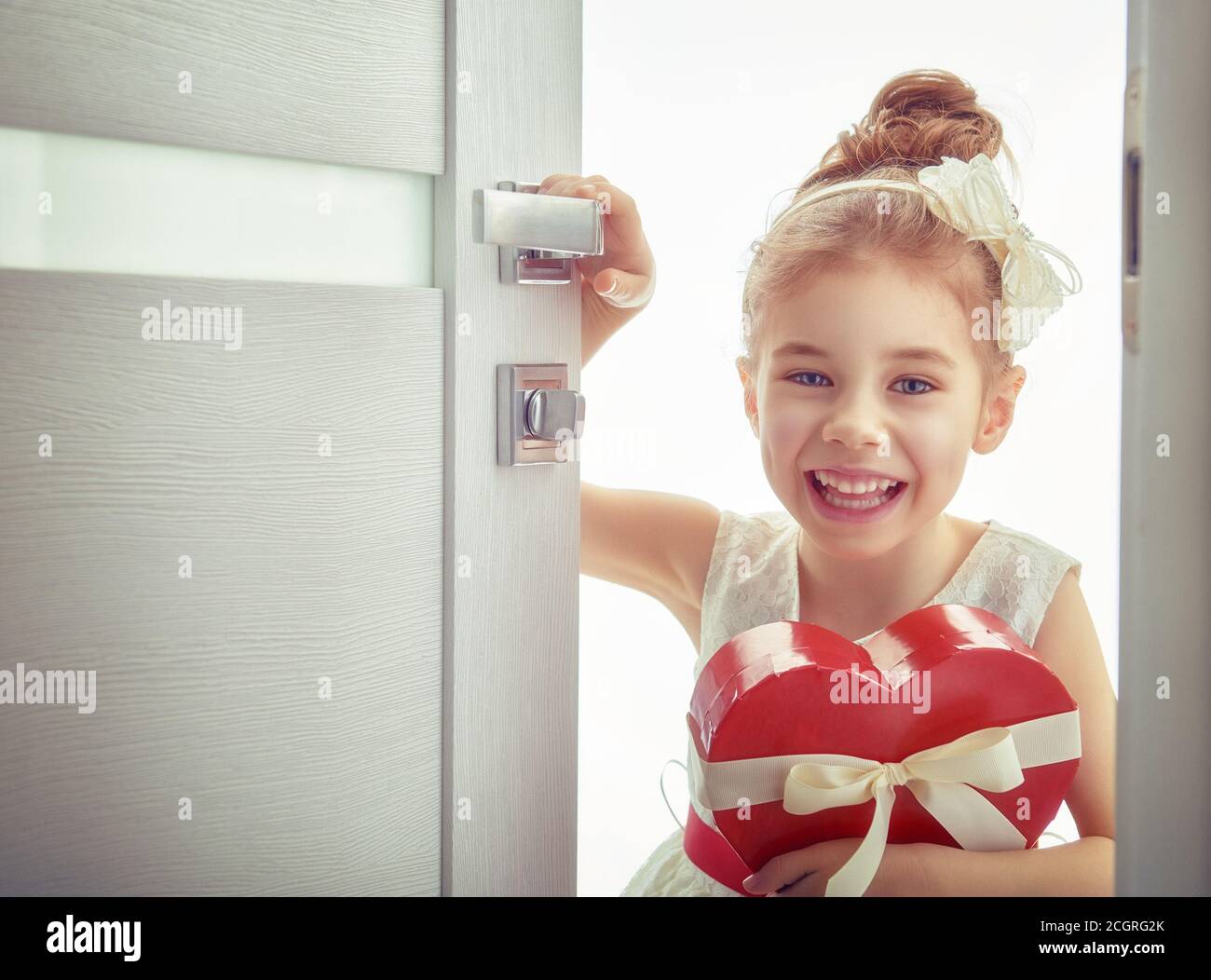 Sweet child girl with red heart. Happy little girl with Valentine's gift opens the door. Wedding, Valentine concept. Stock Photo