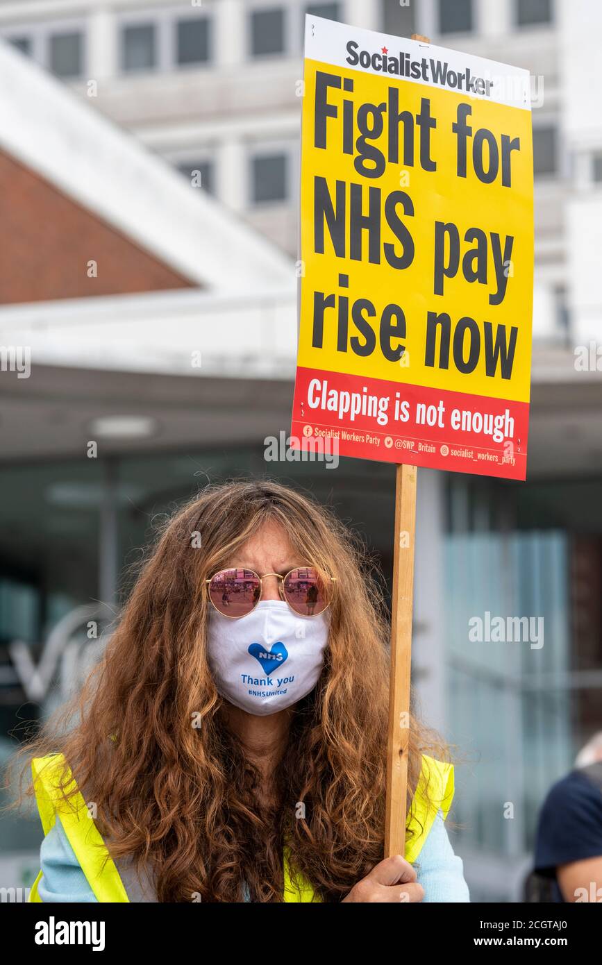 White female protester at a protest against the lack of pay increase for NHS staff. Holding placard with Fight for NHS pay rise now, text. Face mask Stock Photo