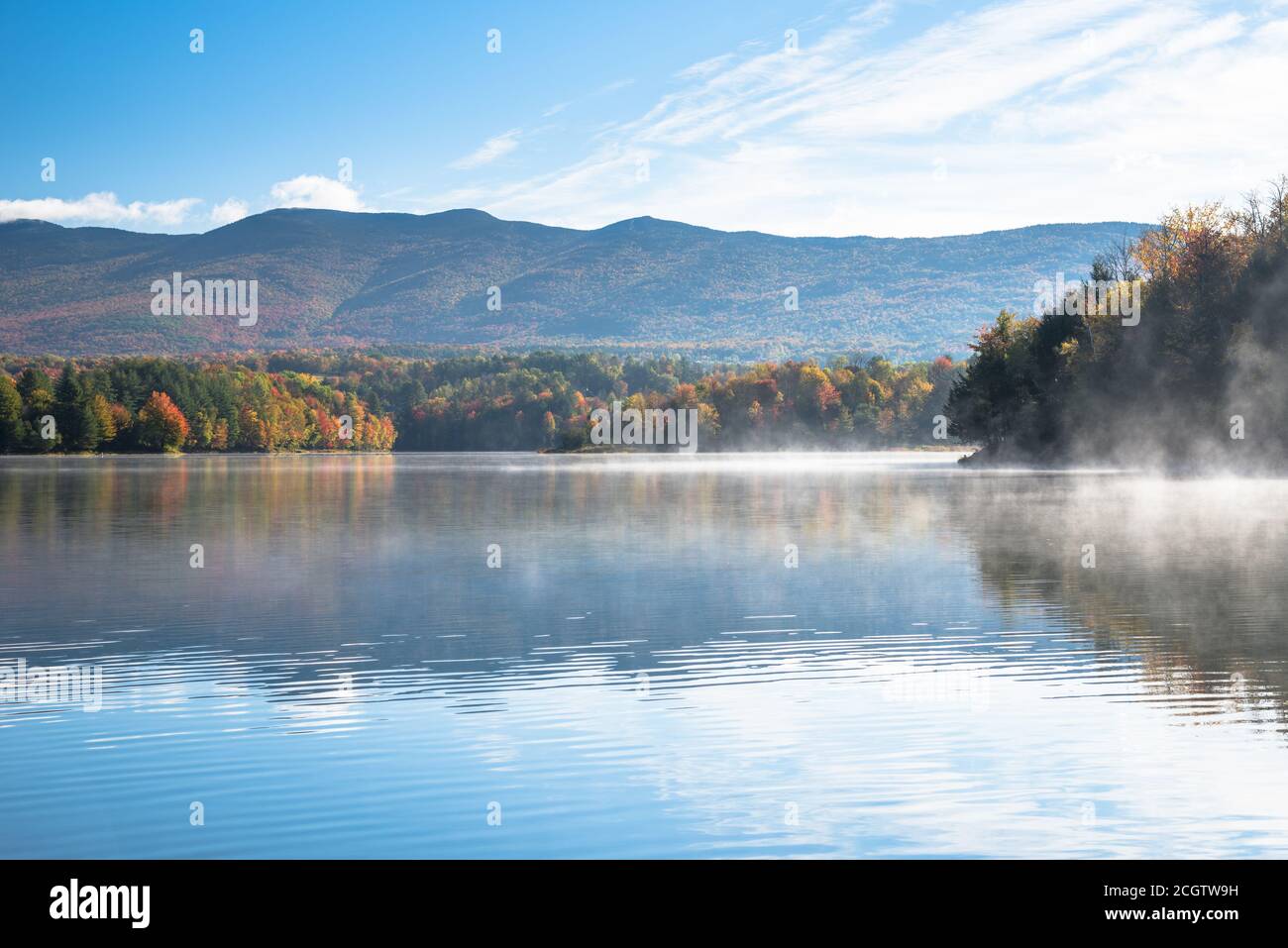 Morning fog drifting over the water of mountain lake with wooded shores at the peak of fall foliage. Waterbury, VT, USA. Stock Photo