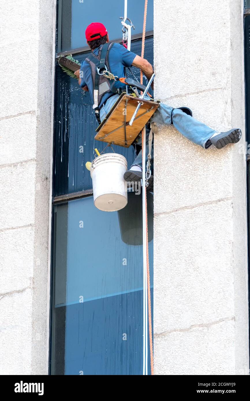 An unidentified window washer working high up on a building. He sits on a very small platform suspended by rope and washes a windows with a large brus Stock Photo