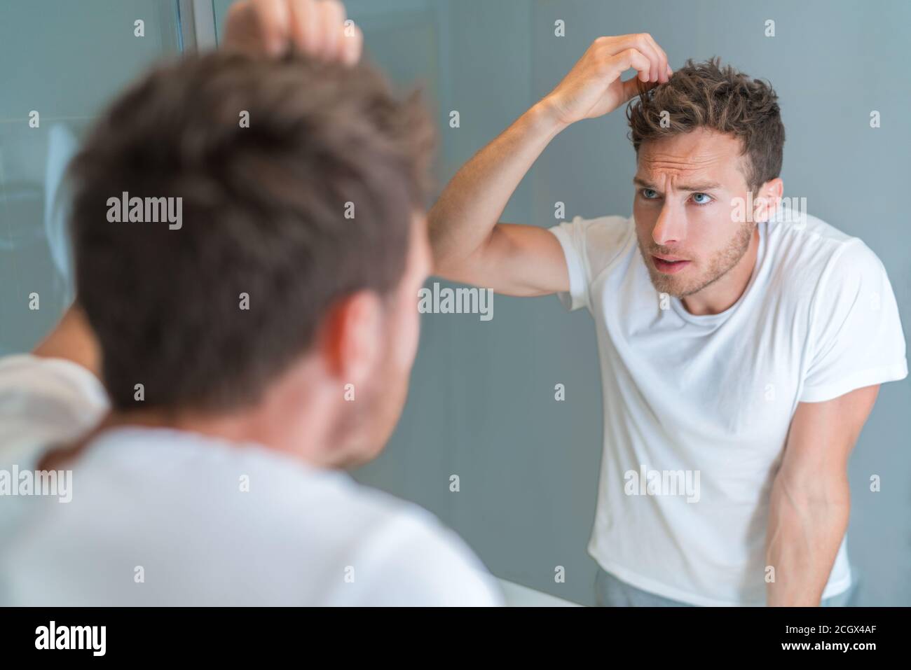 Hair loss man looking in bathroom mirror styling hairstyle with gel or checking for hair loss or grey hairs. Unhappy male health problem Stock Photo