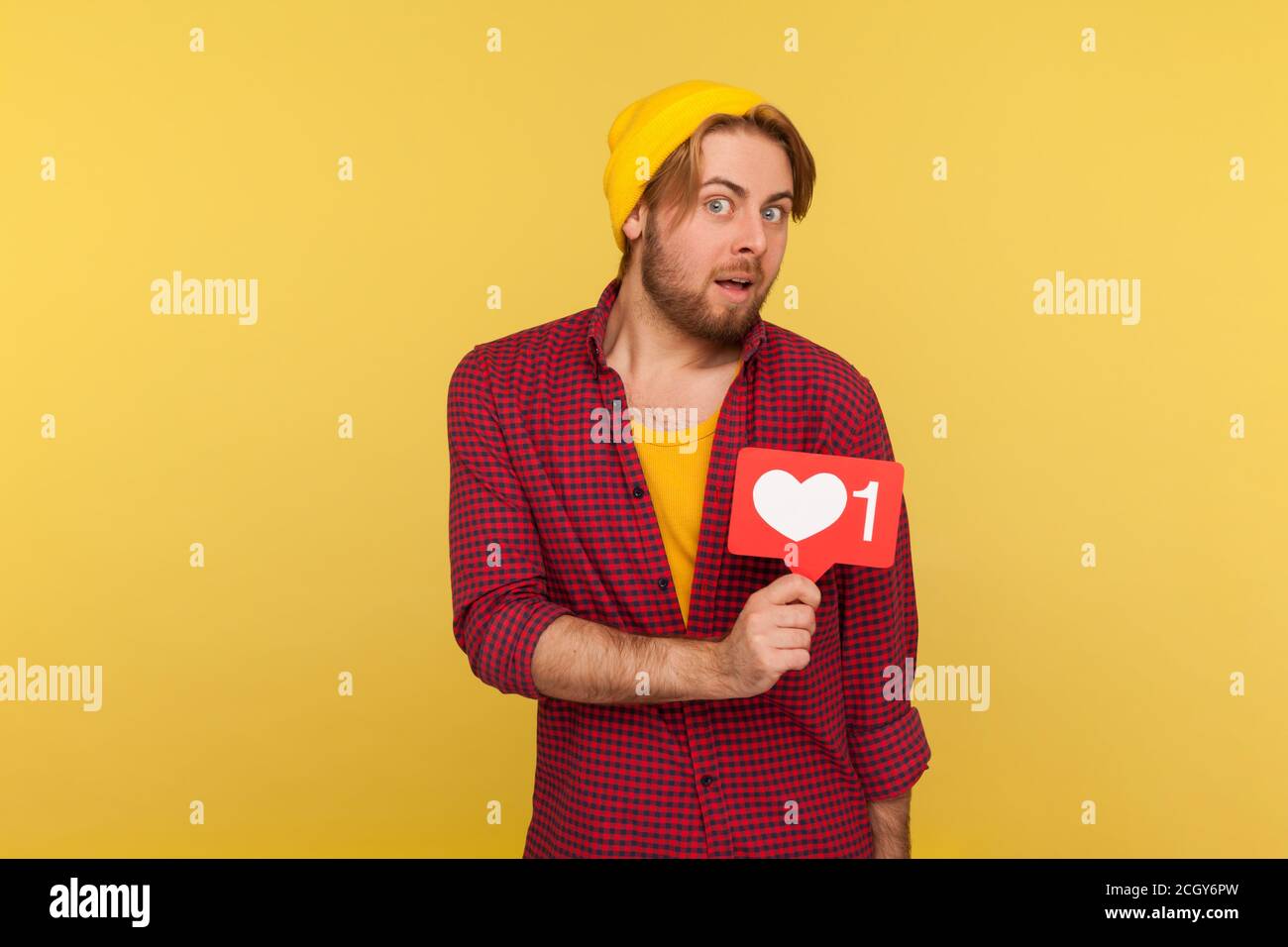 Amazed hipster guy in checkered shirt holding Heart Like icon, counter button of internet popularity in social network, looking at camera big eyes sur Stock Photo
