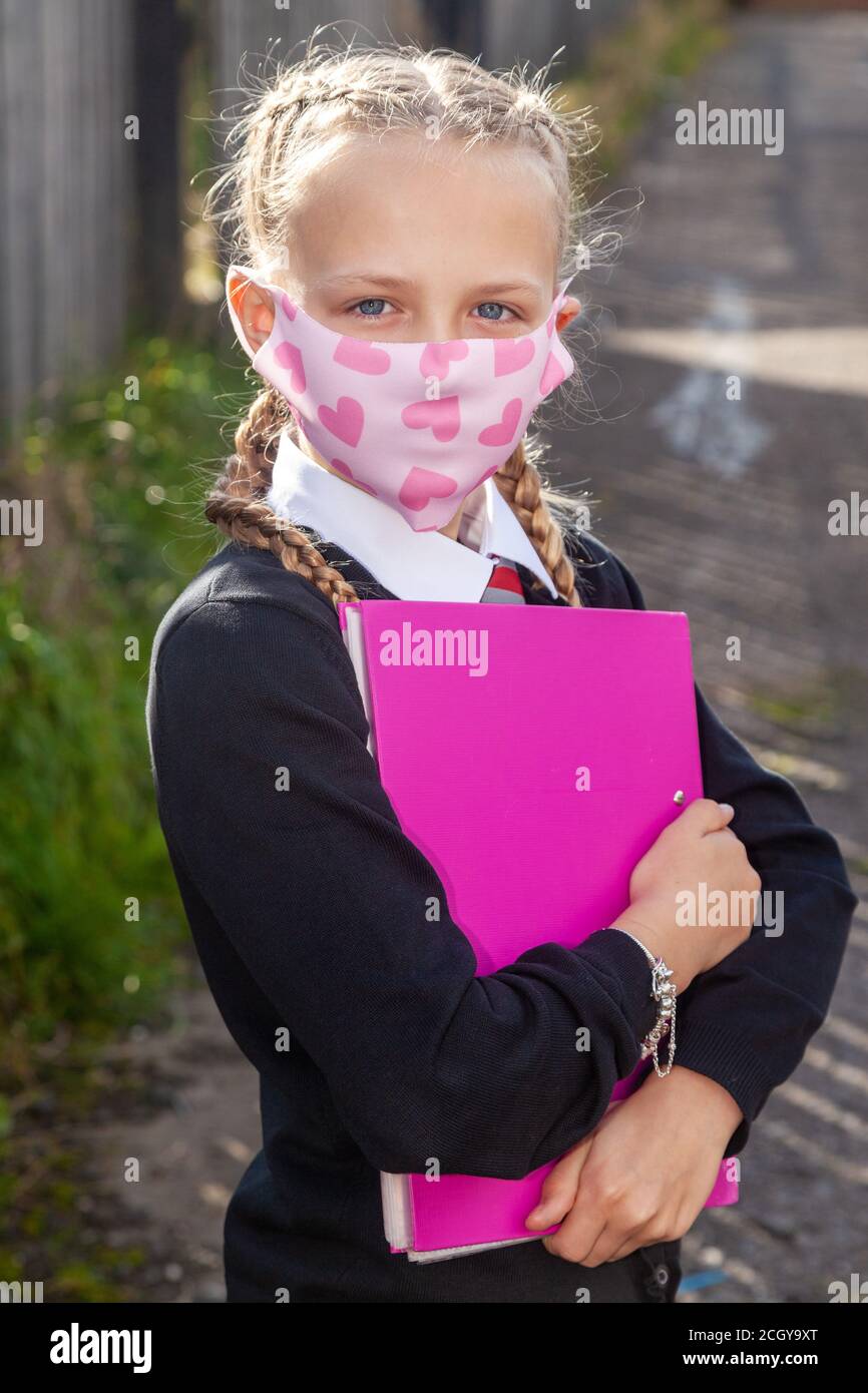 A ten year old schoolgirl with her hair in braids holding a pink A4 folder and wearing a face mask. Stock Photo