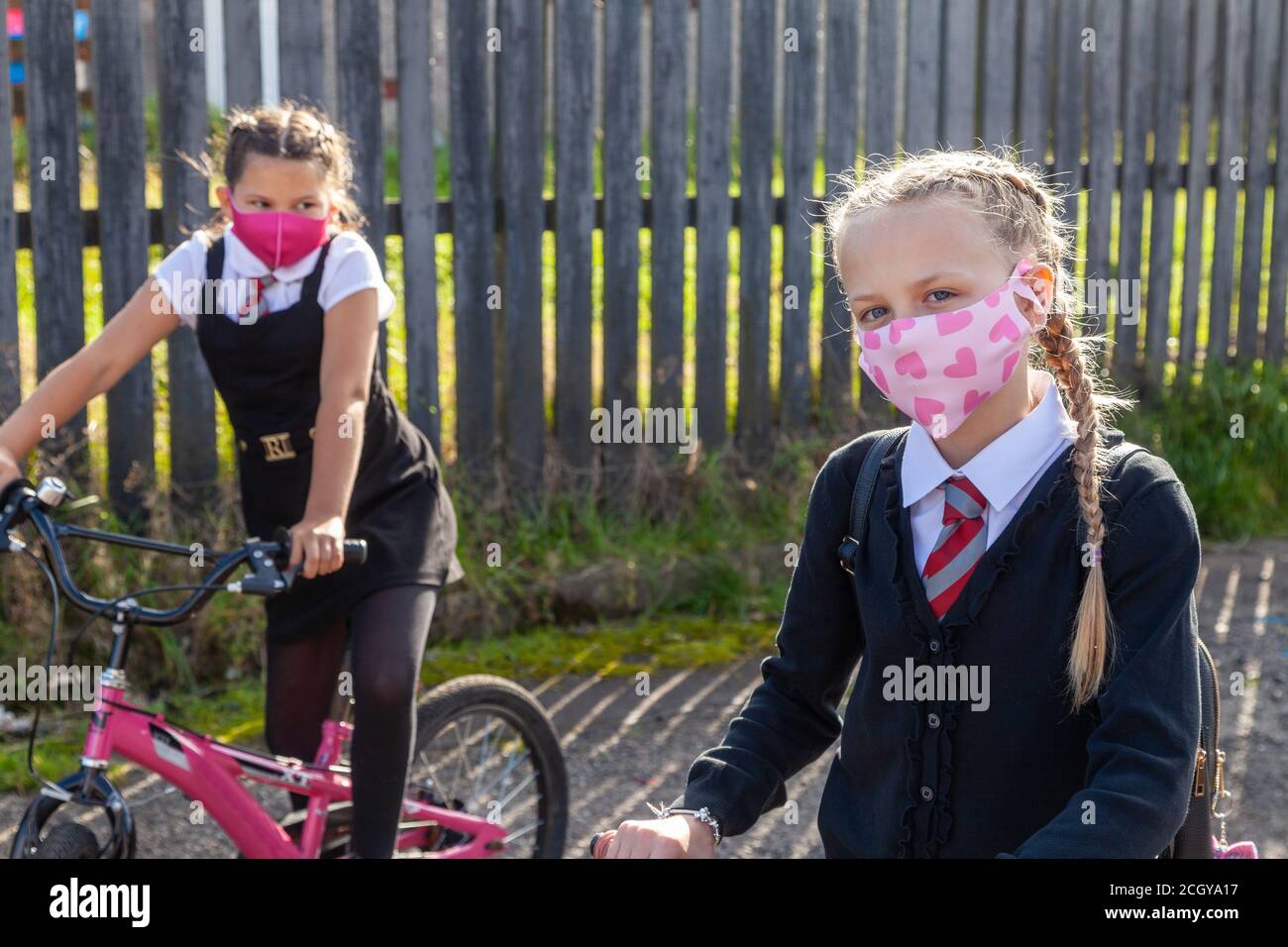 Two ten year old school friends in school uniforms sitting on bicycles and wearing face masks. Stock Photo