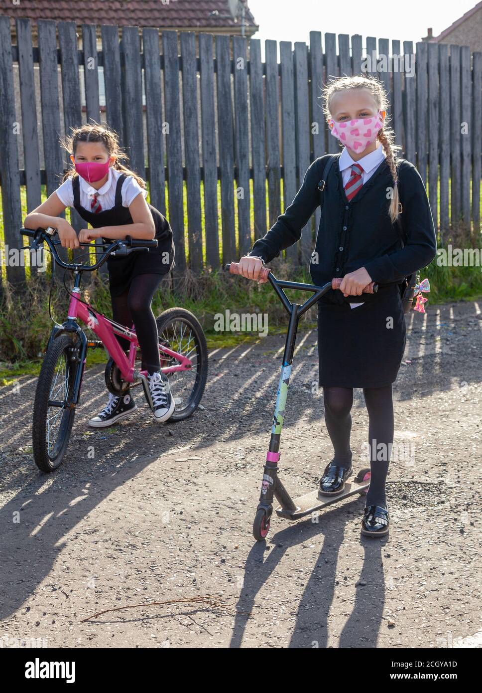 Two ten year old schoolgirls in school uniform one on a bike and the other on a scooter both wearing face masks. Stock Photo